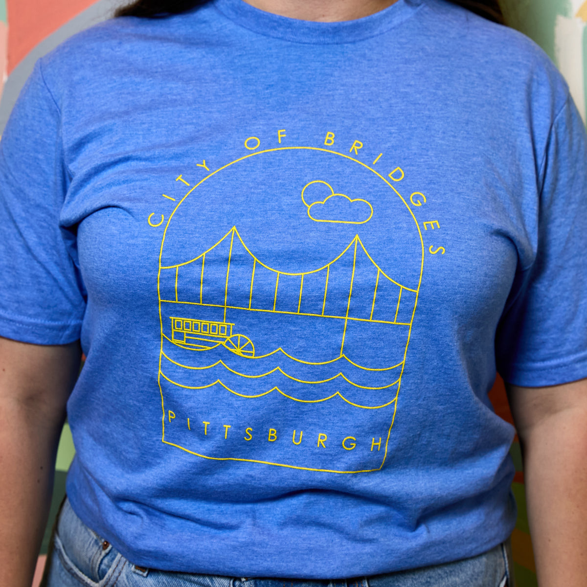 A person wearing a heather blue short-sleeve t-shirt with a yellow line art graphic of the Pittsburgh City of Bridges, paired with blue jeans.