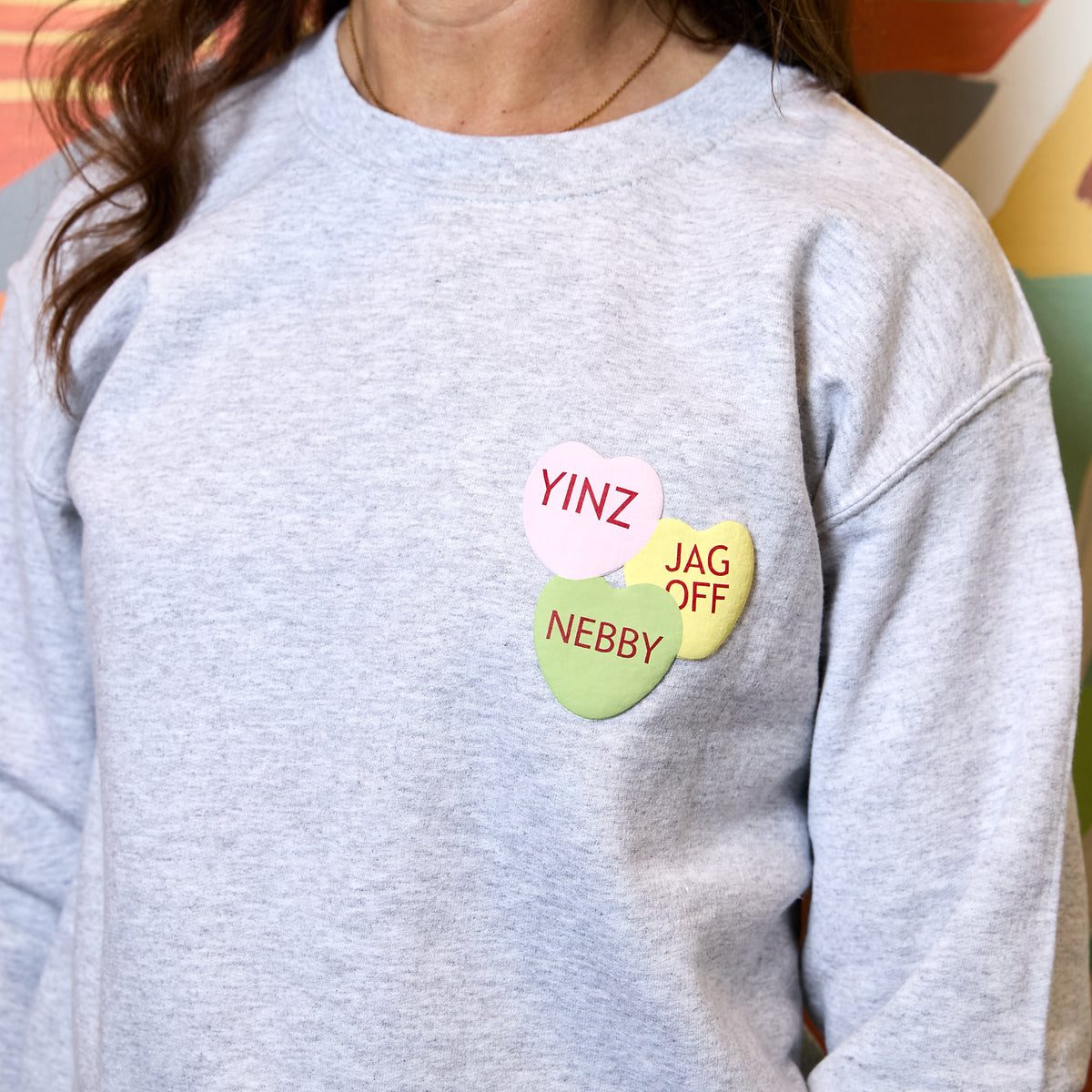 A person wearing a light heather gray sweatshirt with three pastel candy-heart style pins on the chest reading YINZ, JAG OFF, and NEBBY.
