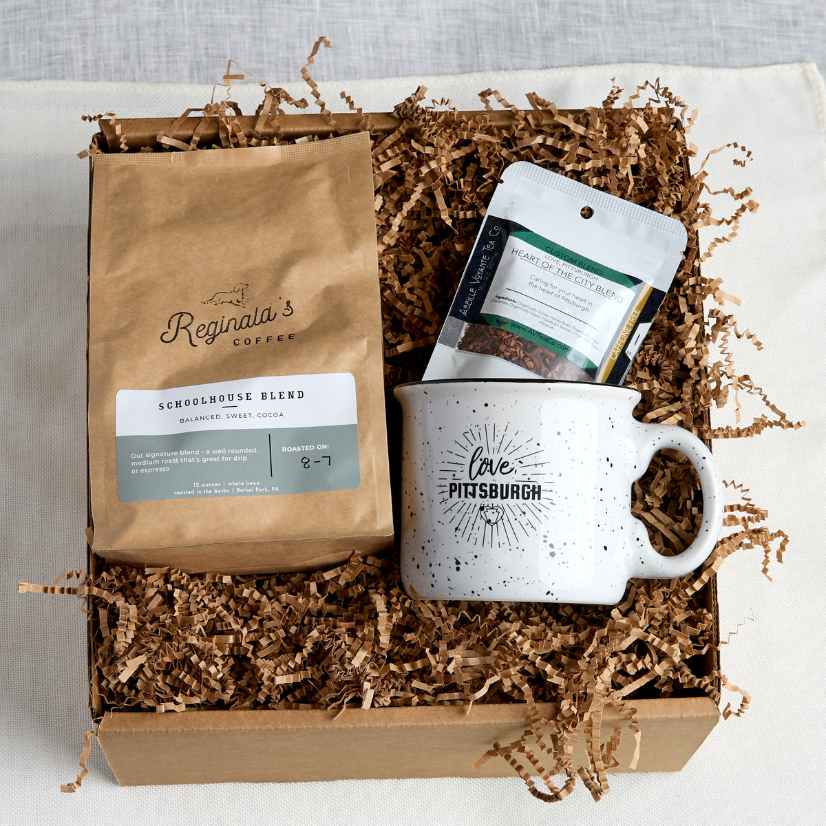 A coffee gift box set containing a bag of Reginald's Coffee Schoolhouse Blend, a white speckled ceramic mug with 'Love Pittsburgh' printed on it, and a package of Abeille Voyante Co. Heart Of The City Blend tea, all arranged in a cardboard box filled with brown paper shred.