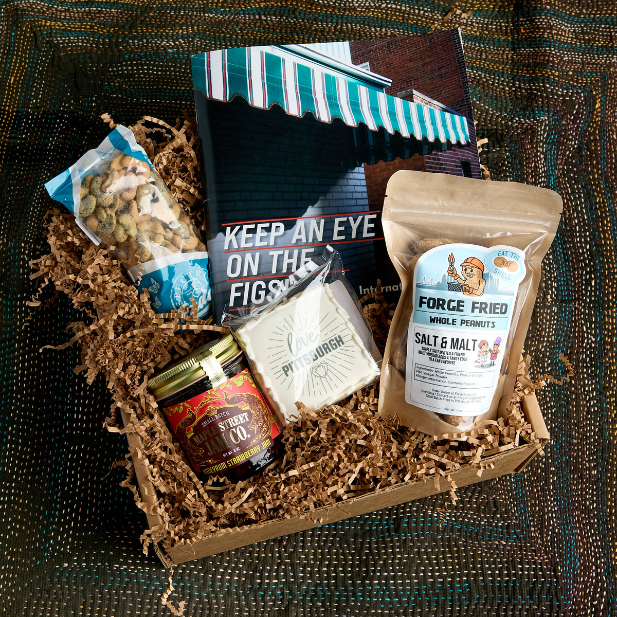 A curated gift box filled with assorted snacks and treats, including a bag of dill cashews, a jar of Maple Street Jam, a packaged logo cookie, a bag of Forge Fried whole peanuts, and a book titled 'Keep an Eye on the Figs,' arranged in brown paper shred on a dark woven blanket.