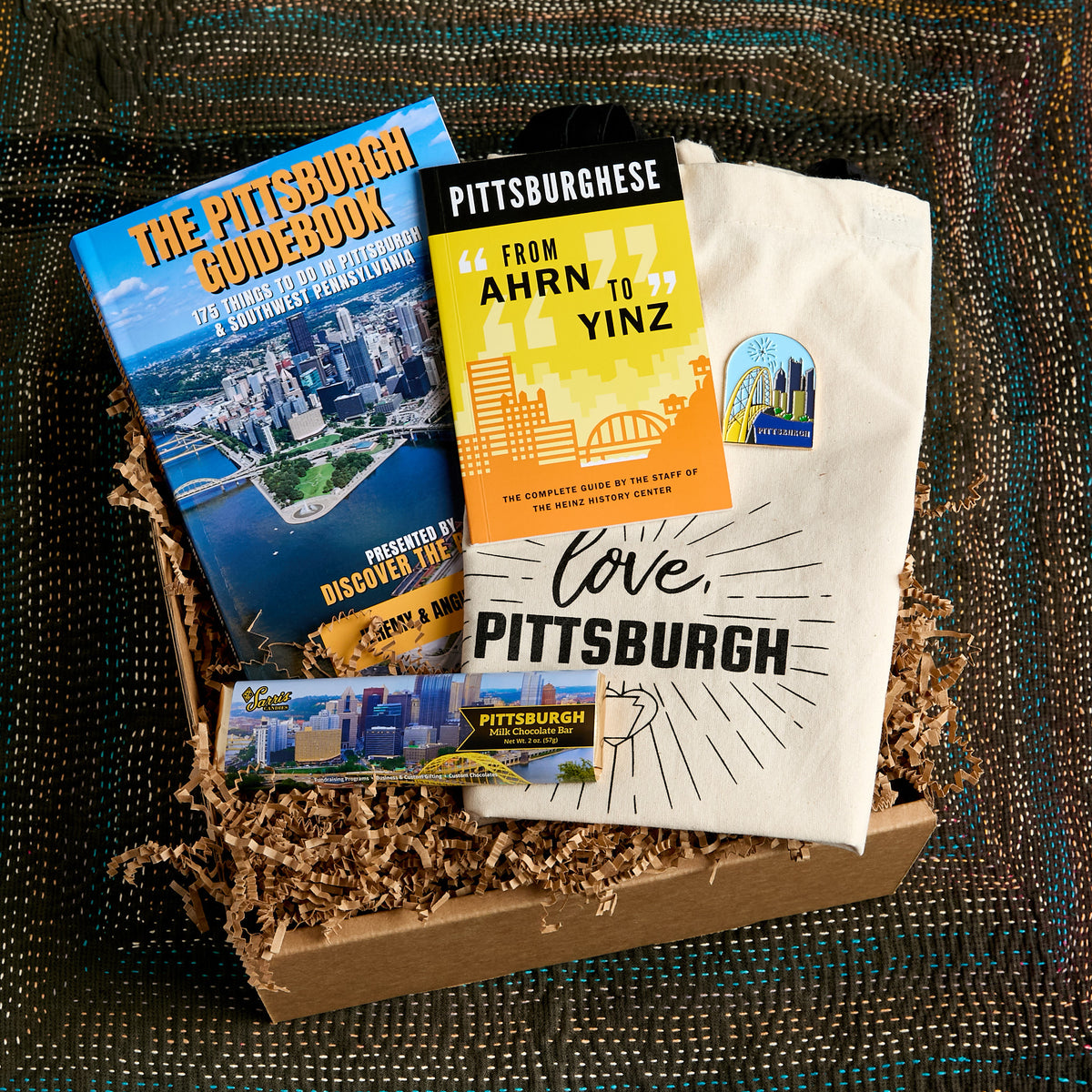 A curated gift box containing a blue 'The Pittsburgh Guidebook', a yellow and black 'Pittsburghese' book, and a white tote printed with 'love, PITTSBURGH' and a skyline graphic, arranged in brown paper shred on a dark woven blanket.