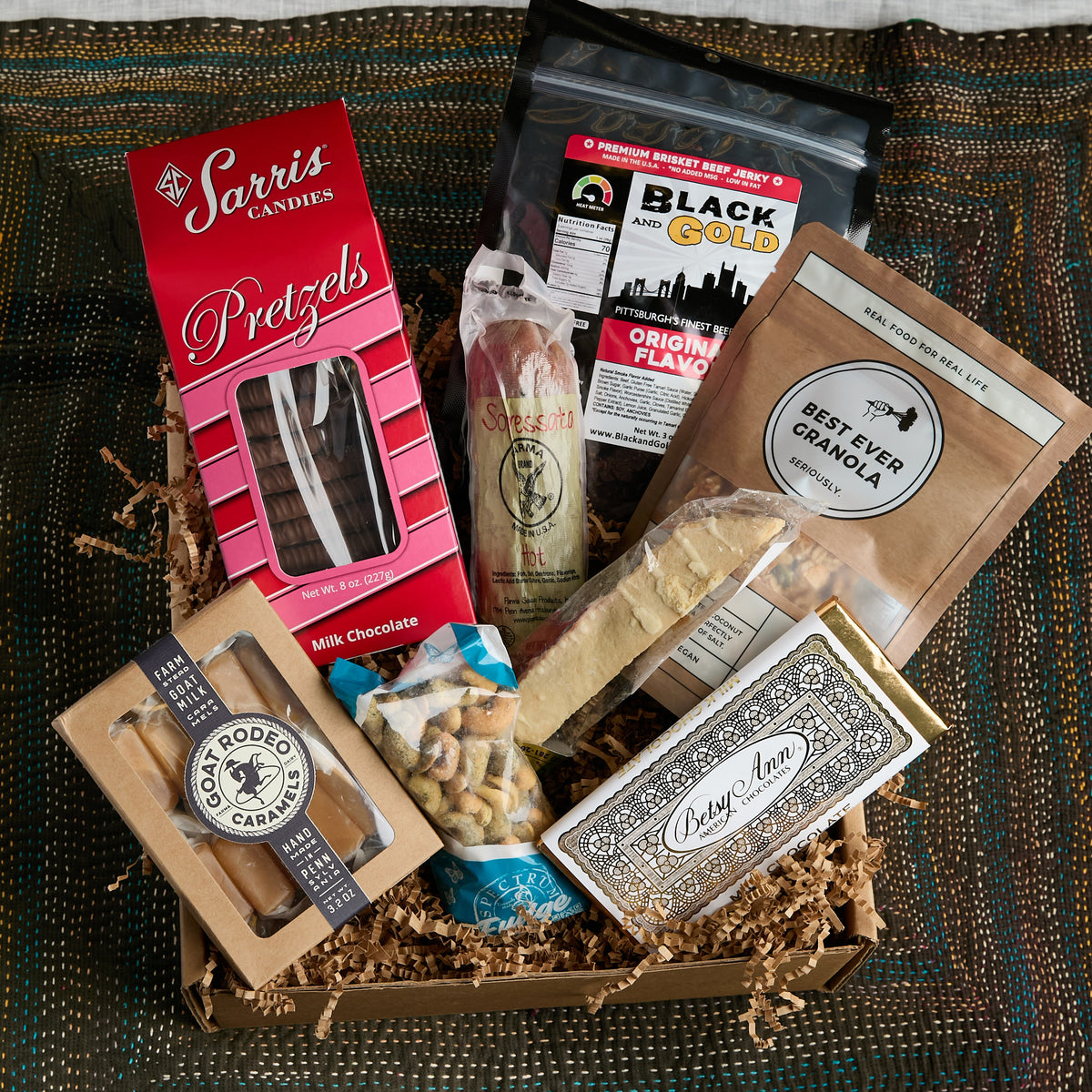A curated gourmet gift box filled with assorted snacks and treats, including Sarris Candies Pretzels, a bag of Black and Gold granola, and other sweet and savory packaged foods, arranged in a cardboard tray with shredded paper on a patterned blanket.
