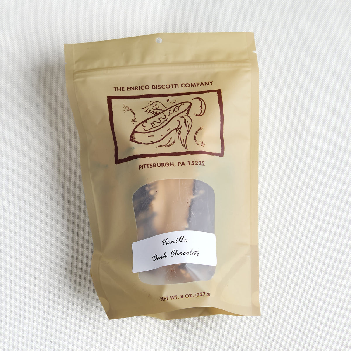 A stand-up pouch of The Enrico Biscotti Company Vanilla Dark Chocolate on a light textured fabric background. The pouch is a warm tan color with a clear window showing the chocolate inside and a white label on the front.
