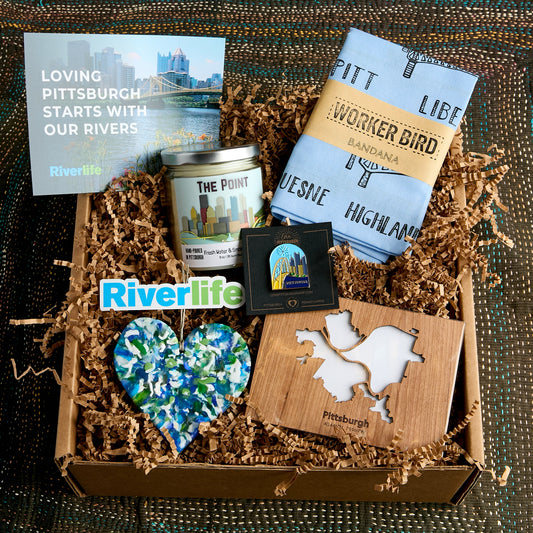 A curated gift box filled with Pittsburgh-themed items: a Riverlife card, a  candle labeled The Point, a folded sky blue bandana with Worker Bird branding, an enamle pin of Pittsburgh, a heart-shaped upcycled plastic heart ornament, and a wooden Pittsburgh map, arranged in brown paper shred.