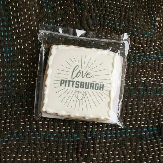 A square white-frosted cookie sealed in a clear plastic bag, placed on a textured dark brown woven blanket. The cookie features the words 'love, PITTSBURGH' in gray lettering with a small heart keystone graphic below.