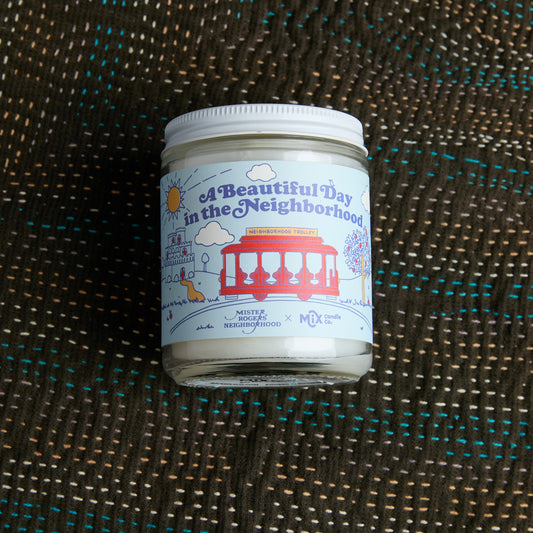 a beautiful day in the neighborhood candle on sky blue label with red trolley and white lid, placed on dark fabric background with colorful stitching