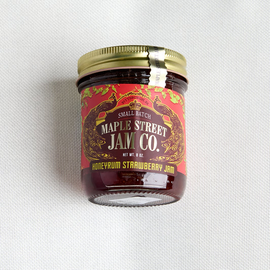 A small glass jar of Maple Street Jam Co. Honeyrum Strawberry Jam with a gold metal lid and a red label, set against a white textured fabric background.