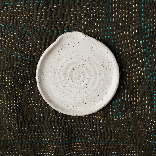 A round, speckled off-white ceramic dish with a small flat edge for resting a spoon, displayed on a dark brown woven textile background.