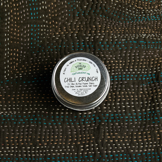 A small round metal tin of Chili Crunch dry rub by Pickled Chef, placed on a dark woven fabric background with visible teal and tan threads.