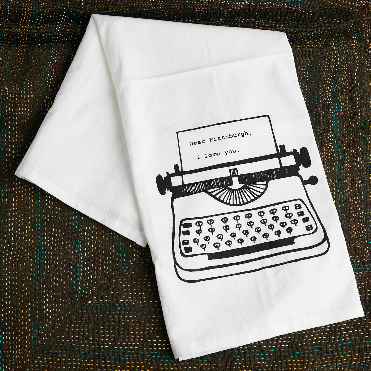 A white kitchen towel with a black line art illustration of a typewriter and the text 'Dear Pittsburgh, I love you.' displayed on a textured brown woven placemat.