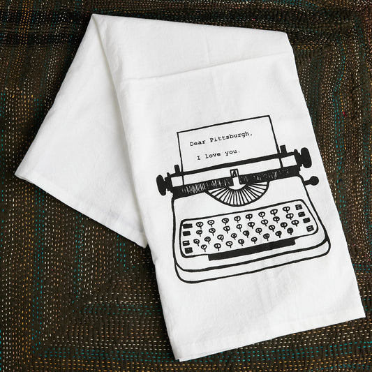 A white kitchen towel with a black line art illustration of a typewriter and the text 'Dear Pittsburgh, I love you.' displayed on a textured brown woven placemat.