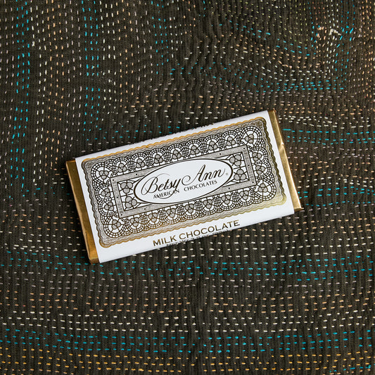 A milk chocolate bar in a white wrapper with ornate black and gold detailing, reading 'Betsy Ann American Chocolate Company' and 'MILK CHOCOLATE,' placed on a textured dark brown woven surface.