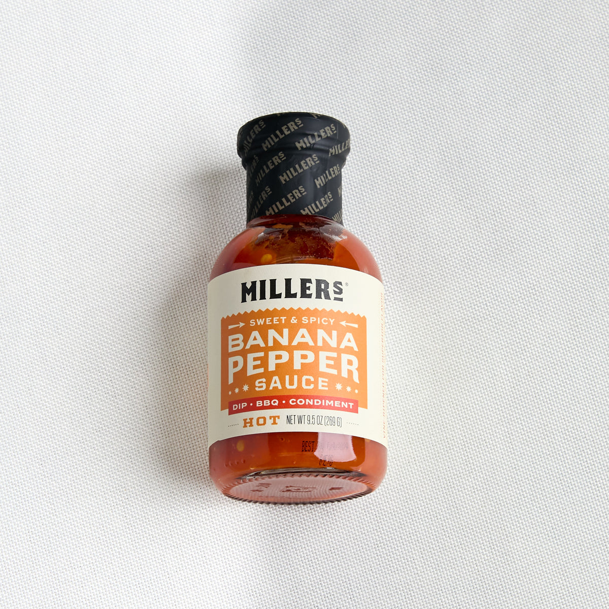 A small bottle of Miller's Banana Pepper Sauce on a textured white fabric background. The bottle is made of amber glass, has a black cap with a black label, and a cream-colored label with orange and black text.