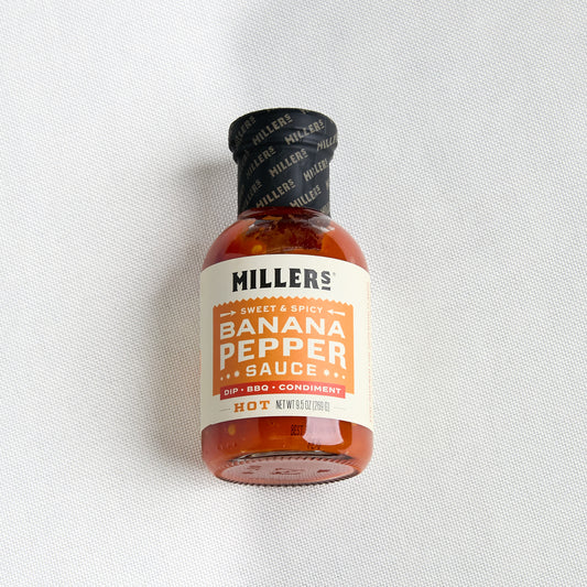 A small bottle of Miller's Banana Pepper Sauce on a textured white fabric background. The bottle is made of amber glass, has a black cap with a black label, and a cream-colored label with orange and black text.