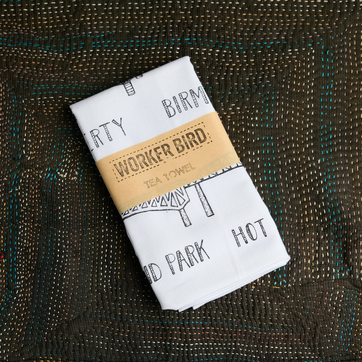 A folded white tea towel with black text and illustration prints, wrapped in a tan paper band reading 'WORKER BIRD TEA TOWEL,' placed on a dark woven surface.