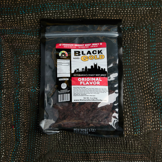A package of Black and Gold Pittsburgh  Beef Jerky, Original Flavor, in a clear plastic bag with a black border and a white, red, and black label, placed on a brown woven mat.
