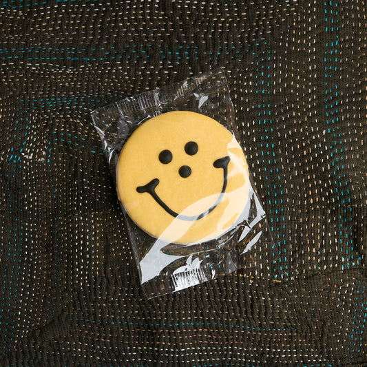 A round yellow smiley face cookie sealed in a clear plastic bag, placed on a dark woven fabric background.