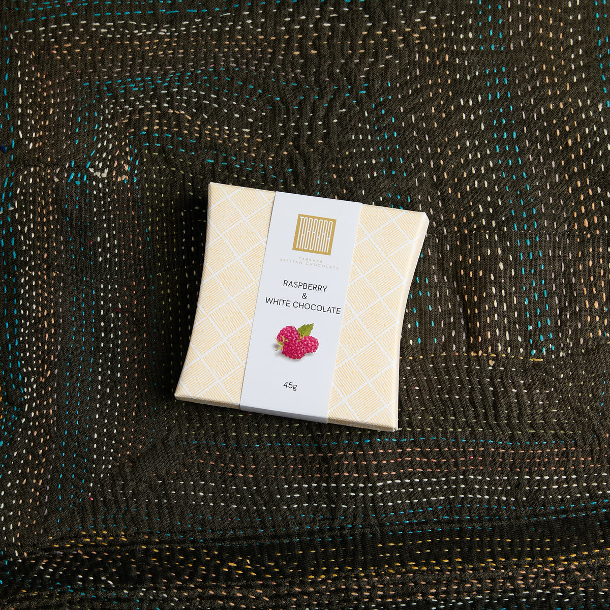 A square box Raspberry + White Chocolate, wrapped in a cream-colored patterned paper sleeve with a white label, placed on a dark brown woven textile background.