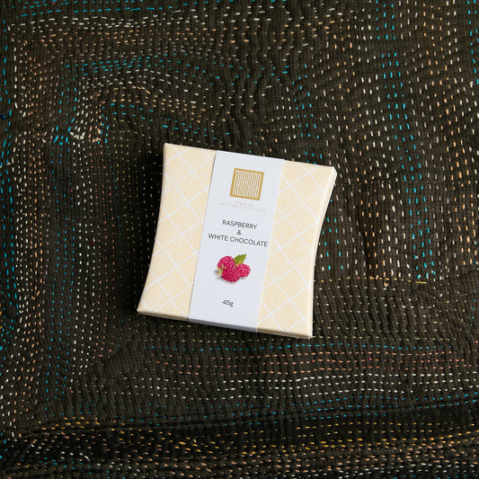 A square box Raspberry + White Chocolate, wrapped in a cream-colored patterned paper sleeve with a white label, placed on a dark brown woven textile background.