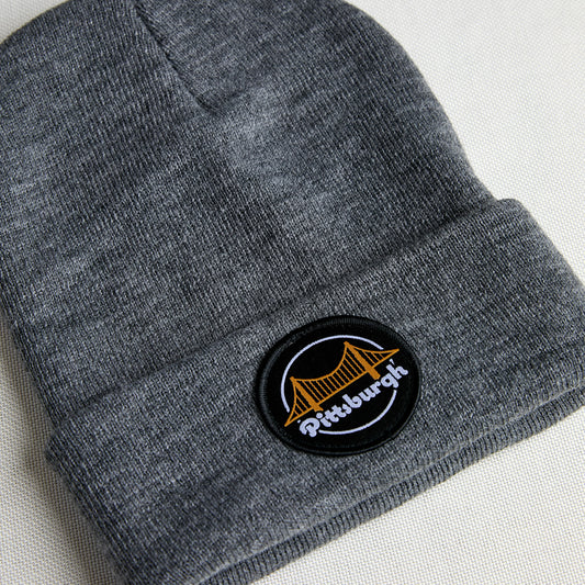 A close-up of a dark gray heather knit beanie with a folded cuff, featuring a circular embroidered patch with a gold bridge and the word 'Pittsburgh' in white on a black background.