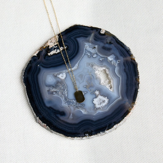 A gold necklace with a small gold palin dog tag pendant, draped over a round agate slice coaster on a textured white fabric background.