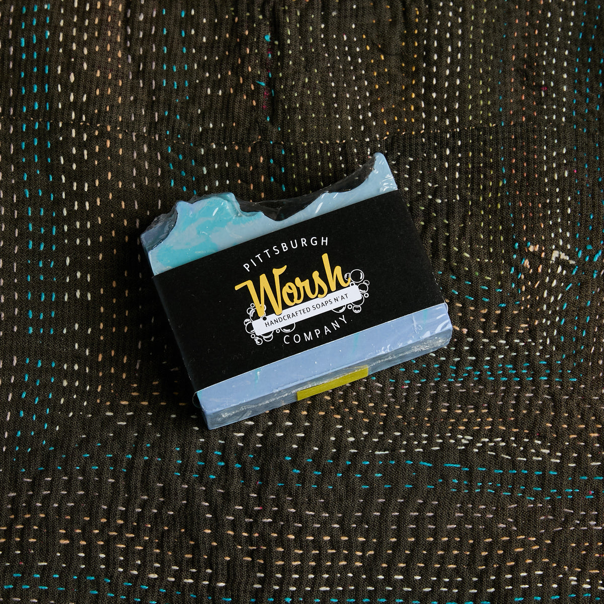 A bar of Pittsburgh Artisan Crafted Soap Bar resting on a textured brown woven blanket. The soap features a black paper wrapper with yellow and white branding, and visible light blue and white marbled soap layers at the top and bottom.
