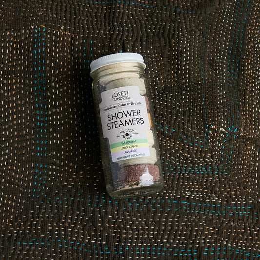 A glass jar of Lovett Sundries Shower Steamers filled with colorful steamer pucks, placed on a dark brown woven blanket with teal stitching.