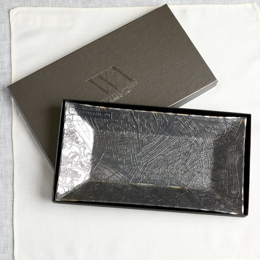 A rectangular aluminum tray with an embossed city map of Pittsburgh pattern, shown open next to its matching dark grey textured presentation box on a white fabric background.