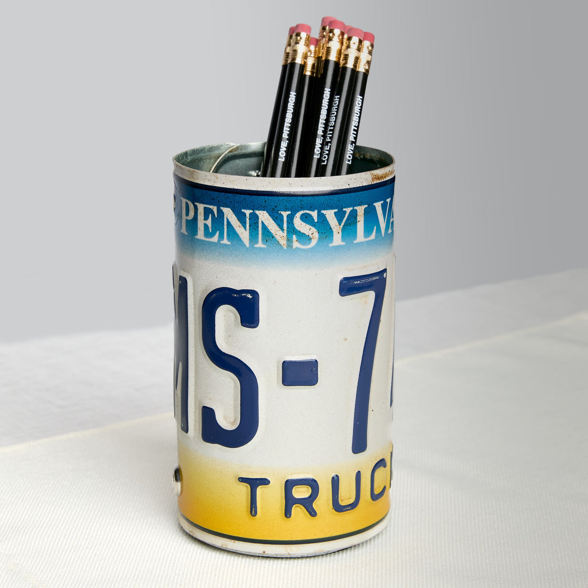 A novelty pencil holder shaped like a vintage Pennsylvania license plate truck plate, filled with four black pencils, set on a white textured surface against a light gray background.