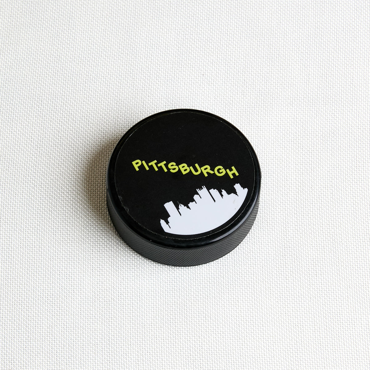 A black circular hockey puck with "Pittsburgh" and skyline, shown from a top-down view on a plain white background.