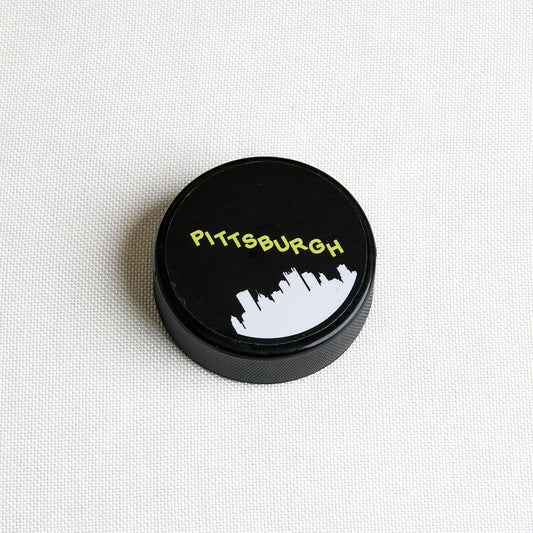 A black circular hockey puck with "Pittsburgh" and skyline, shown from a top-down view on a plain white background.