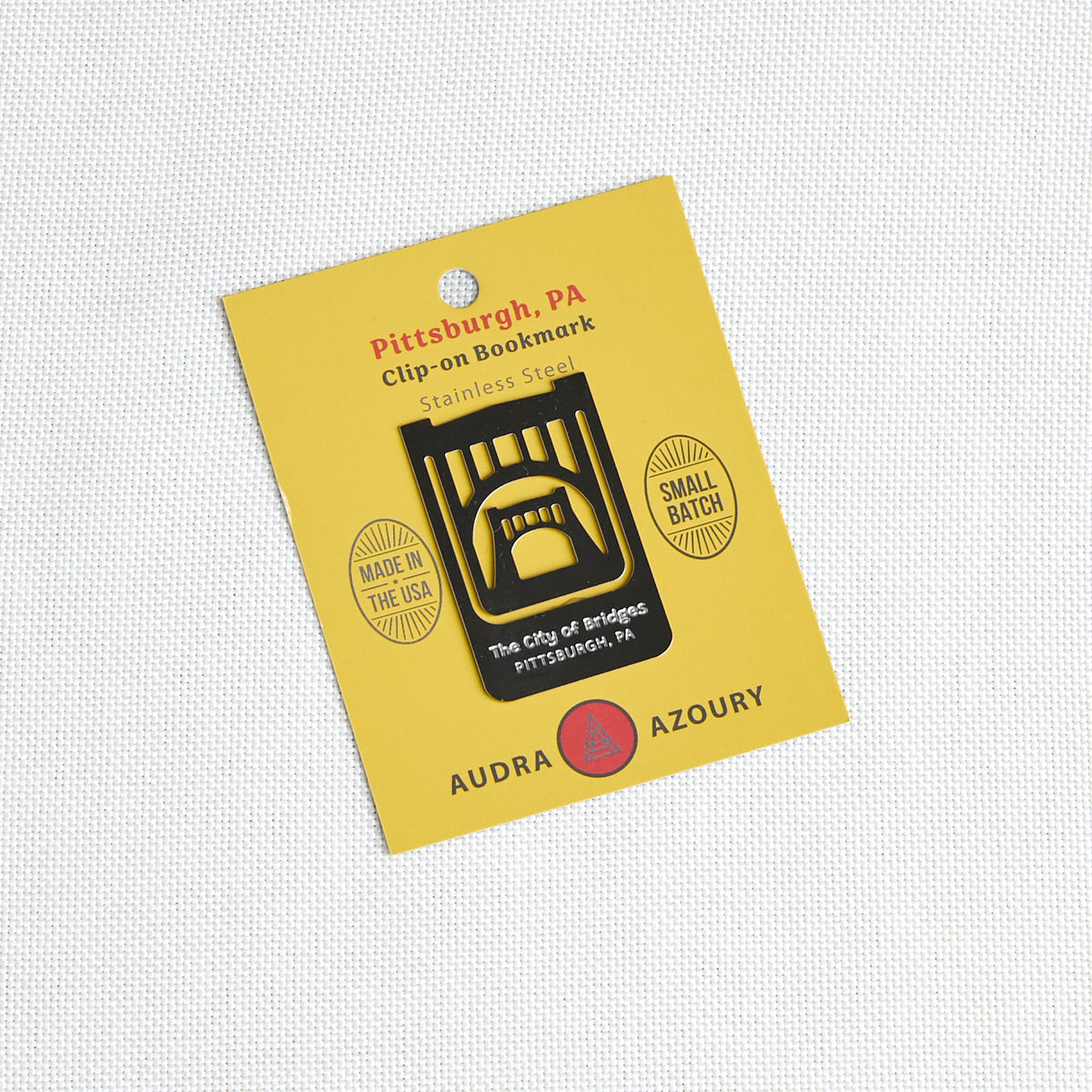 A yellow card backing holding a small clip-on bookmark shaped like a black bridge over water, with printed text reading 'Pittsburgh, PA', 'Clip-on Bookmark', 'Stainless Steel', and 'Audra Azoury'.