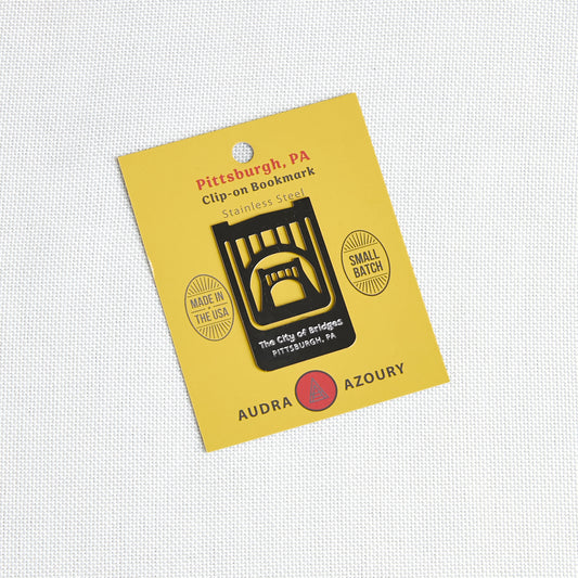 A yellow card backing holding a small clip-on bookmark shaped like a black bridge over water, with printed text reading 'Pittsburgh, PA', 'Clip-on Bookmark', 'Stainless Steel', and 'Audra Azoury'.