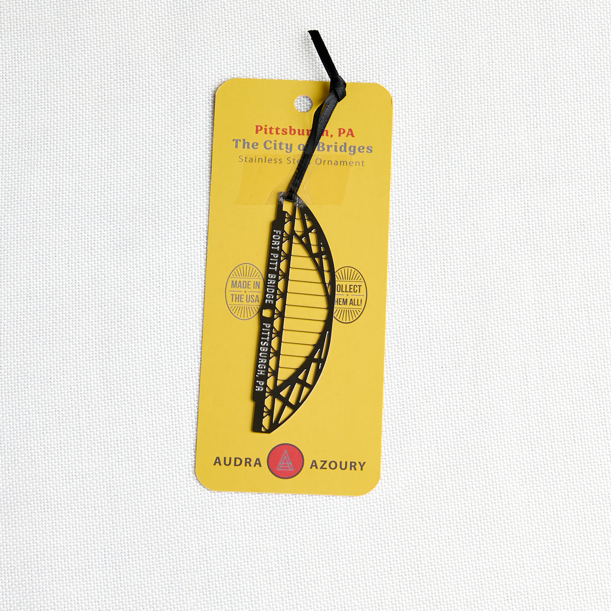 A metal Pittsburgh City Bridges ornament of the Fort Pitt Bridge, attached to a bright yellow backing card, photographed on a white textured fabric background.