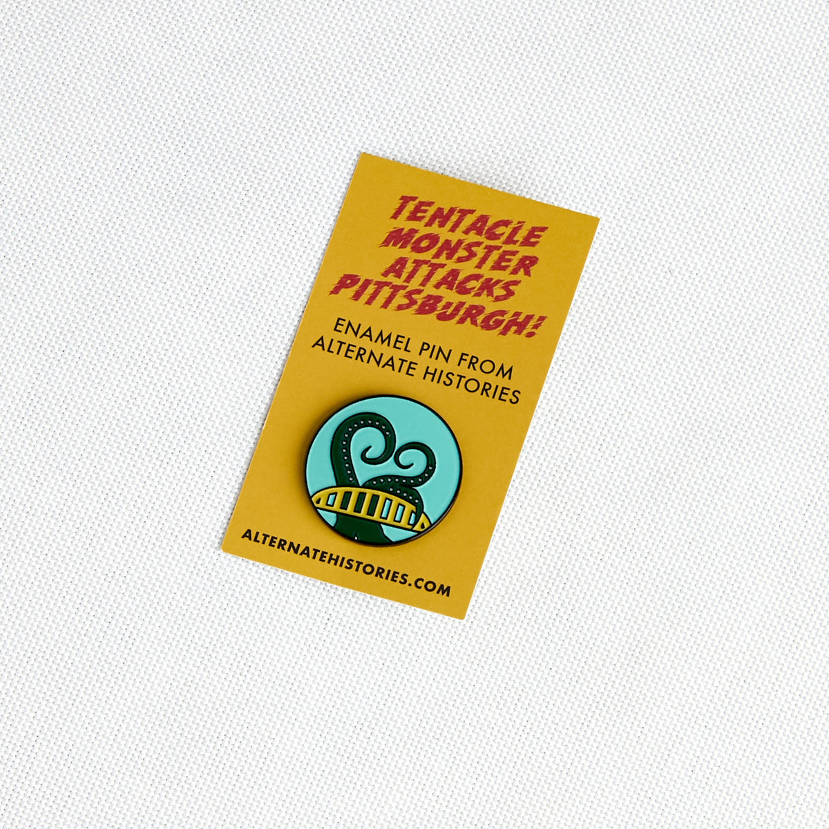 A yellow enamel pin backing card featuring red text that reads 'Tentacle Monster Attacks Pittsburgh!' and a circular pin illustration of a green tentacle monster on a light blue background. The card also includes the text 'ENAMEL PIN FROM ALTERNATE HISTORIES' and 'ALTERNATEHISTORIES.