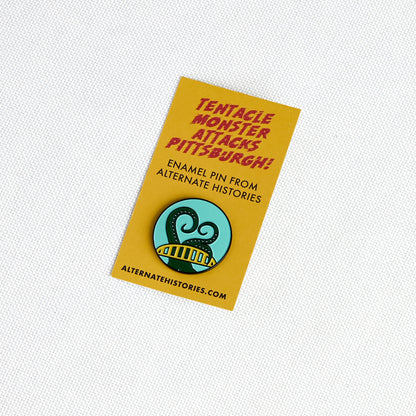 A yellow enamel pin backing card featuring red text that reads 'Tentacle Monster Attacks Pittsburgh!' and a circular pin illustration of a green tentacle monster on a light blue background. The card also includes the text 'ENAMEL PIN FROM ALTERNATE HISTORIES' and 'ALTERNATEHISTORIES.