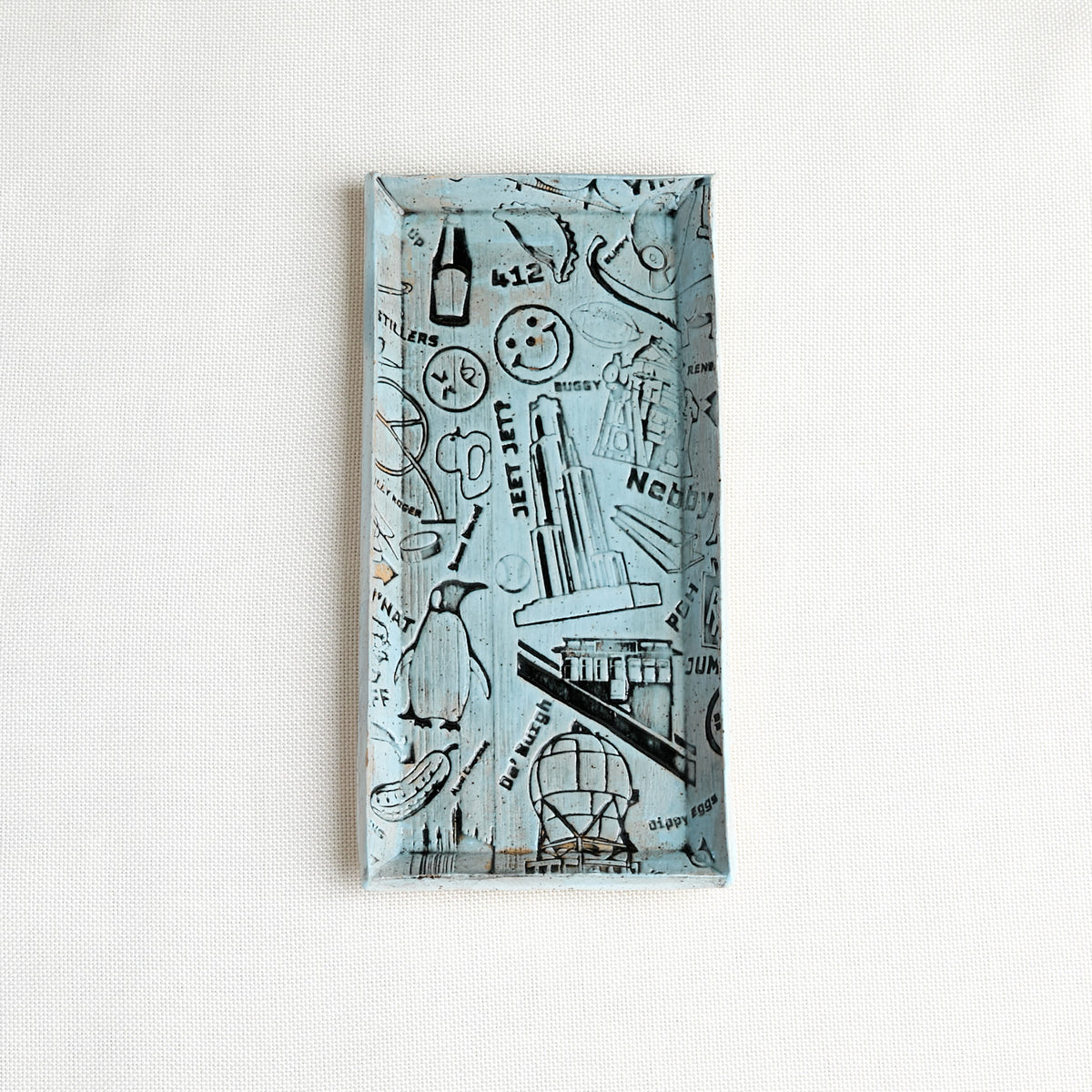A rectangular decorative tray with a light blue background and iconic Pittsburgh illustrations, photographed against a white textured wall.