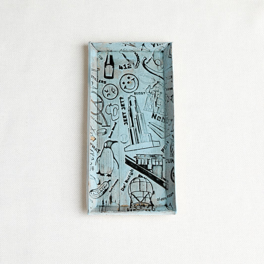 A rectangular decorative tray with a light blue background and iconic Pittsburgh illustrations, photographed against a white textured wall.