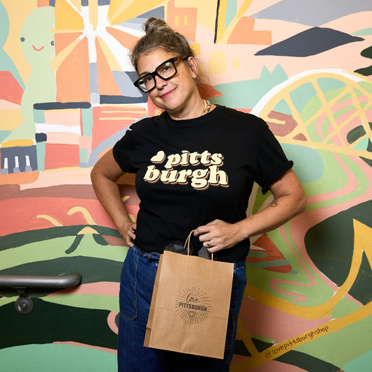 A woman wearing black glasses and a black t-shirt with 'pitts burgh' printed in cream and green, holding a brown paper bag with the 'Pittsburgh Art Museum' logo, standing in front of a colorful painted mural.