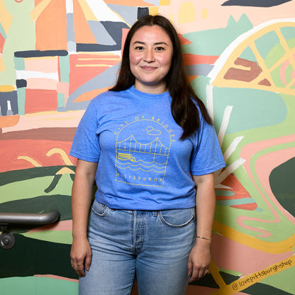 A woman wearing a blue crewneck t-shirt with a yellow outline graphic of a bridge and the words 'PITTSBURGH, PA' and 'City Of Bridges', standing in front of a colorful painted mural.