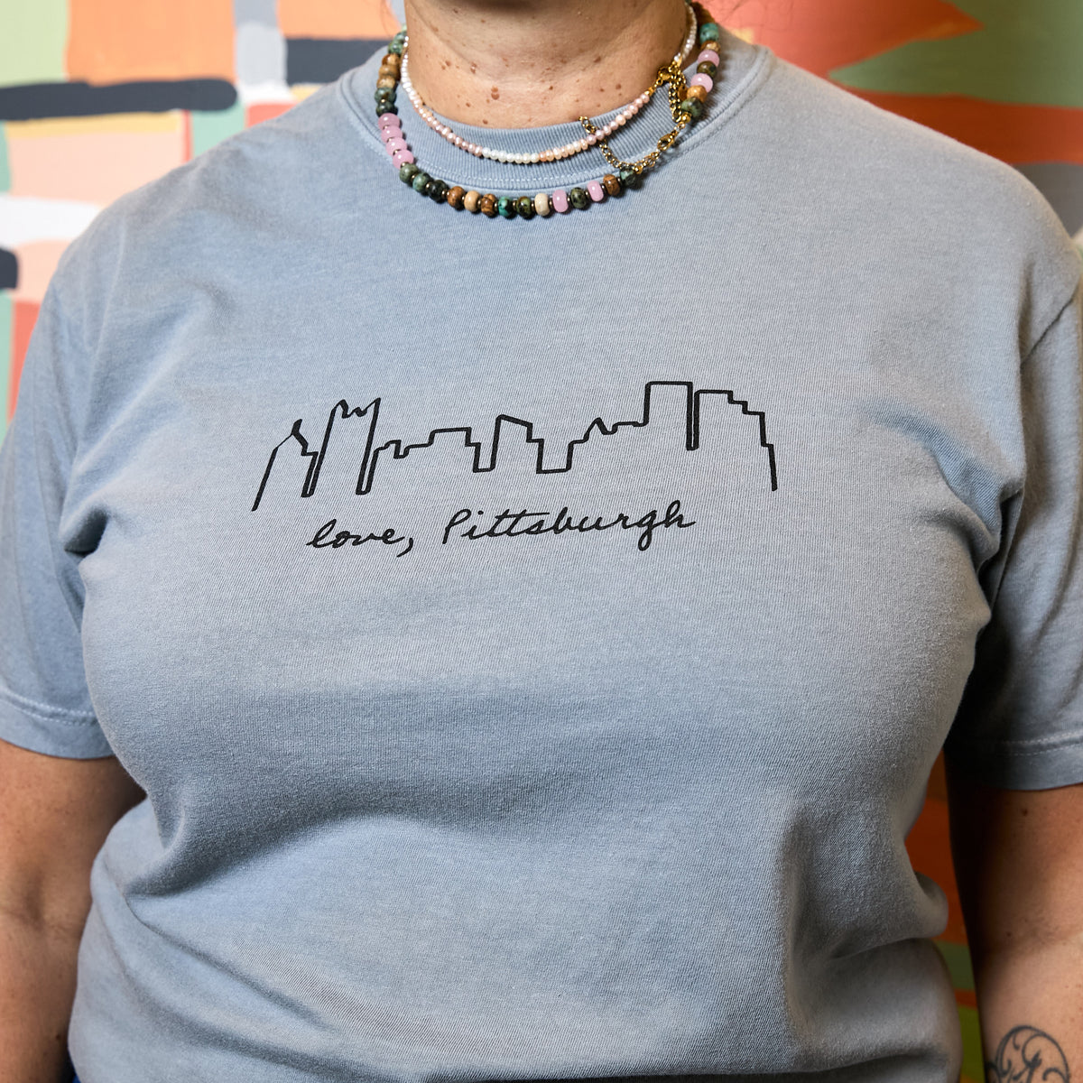 A person wearing a light blue crewneck t-shirt with a black outline of the Pittsburgh skyline and the text 'love, Pittsburgh' printed on the chest. The person is also wearing layered beaded necklaces.