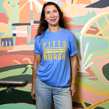 A woman wearing a blue crewneck t-shirt with 'PITTSBURGH' printed in yellow across the chest, standing in front of a colorful abstract mural.