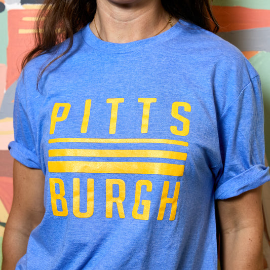 A person wearing a heather blue short-sleeve T-shirt with rolled sleeves and a large yellow 'PITTSBURGH' graphic across the chest, photographed in a real indoor setting with a colorful wall in the background.