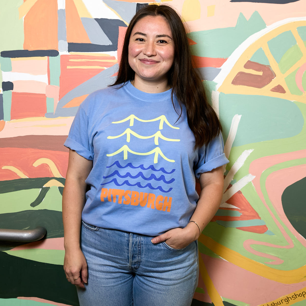 A woman wearing a light blue graphic t-shirt with a Pittsburgh skyline design, standing in front of a colorful abstract mural.