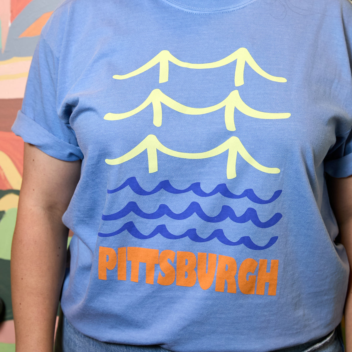 A person wearing a light blue graphic t-shirt featuring a yellow bridge design, blue waves, and the word 'PITTSBURGH' in orange.