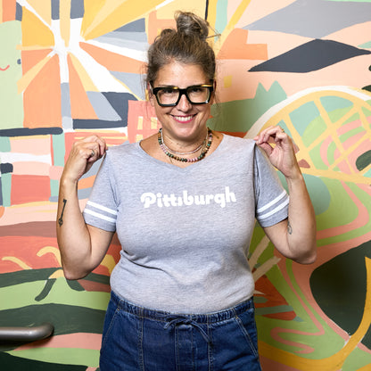 A woman wearing a heather gray t-shirt with 'Pittsburgh' printed in white across the chest, posing in front of a colorful abstract mural.