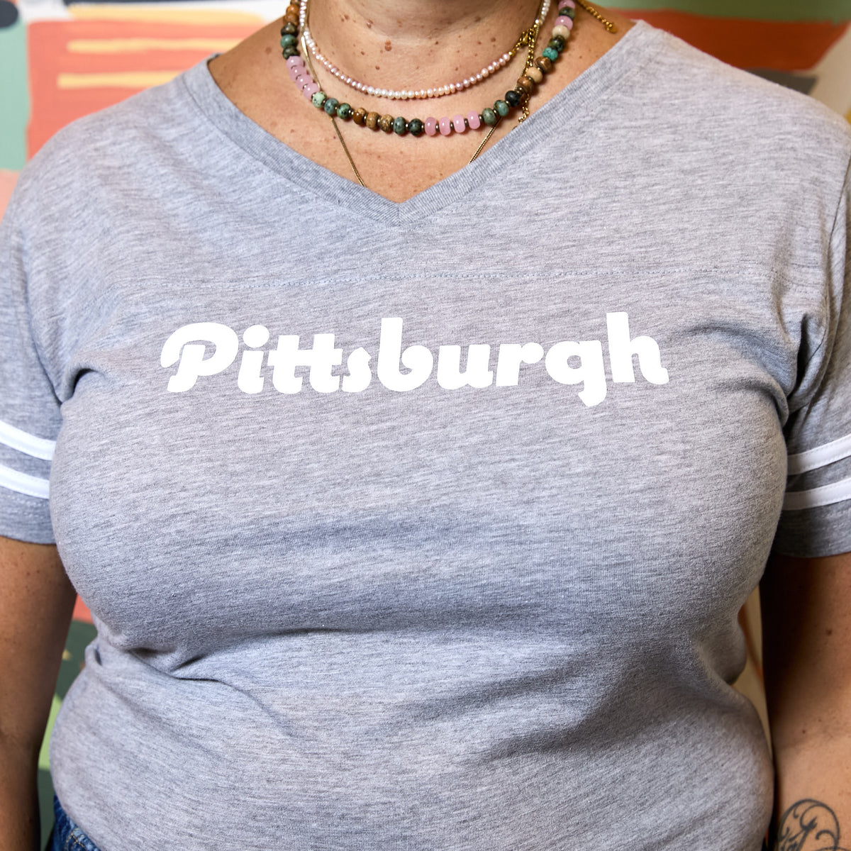 A person wearing a heather gray V-neck T-shirt with the word "Pittsburgh" printed in large white letters across the chest and white stripes on the sleeves.