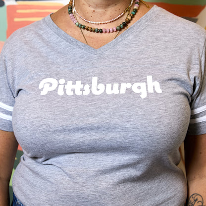 A person wearing a heather gray V-neck T-shirt with the word "Pittsburgh" printed in large white letters across the chest and white stripes on the sleeves.