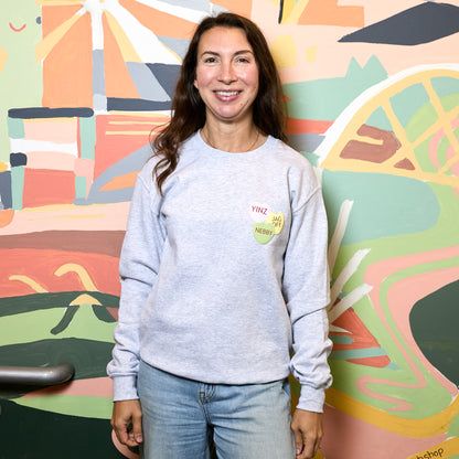 A woman smiling and wearing a light gray crewneck sweatshirt with a colorful heart graphic on the chest, paired with blue jeans, standing in front of a vibrant, multicolored abstract mural.