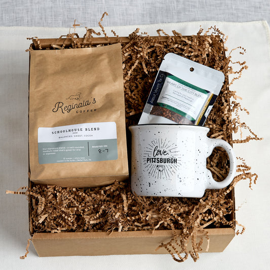 A coffee gift box set containing a bag of Reginald's Coffee Schoolhouse Blend, a white speckled ceramic mug with 'Love Pittsburgh' printed on it, and a package of Abeille Voyante Co. Heart Of The City Blend tea, all arranged in a cardboard box filled with brown paper shred.
